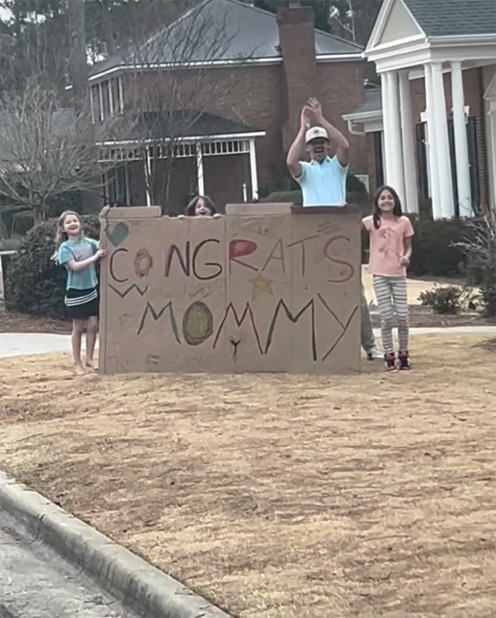Three little girls and their father clap behind a sign that reads 'Congrats Mommy.'