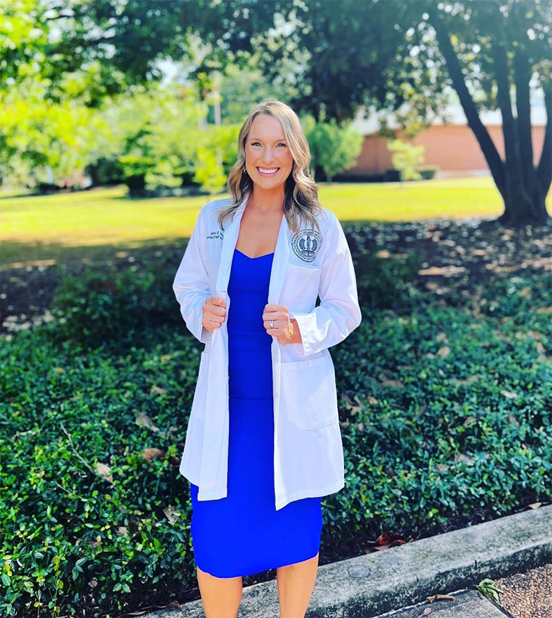 A woman wearing a lab coat stands outside and smiles at the camera.