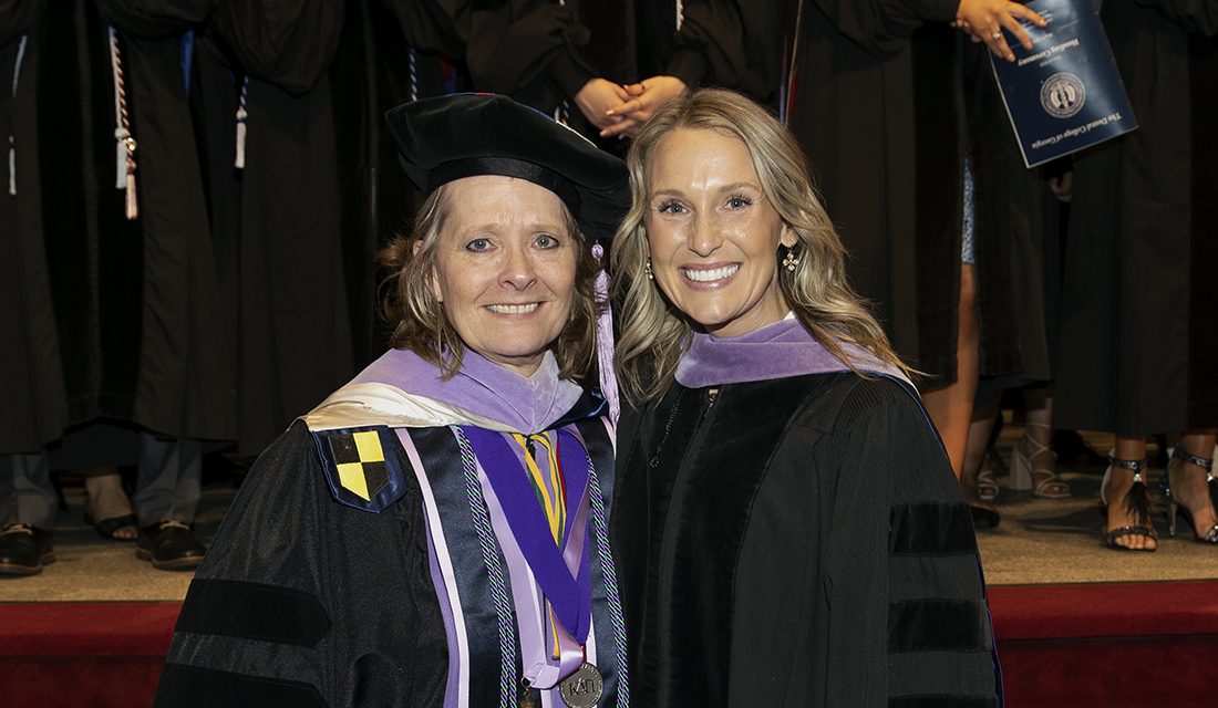 A woman wearing graduation regalia, including doctoral hood, shares a smile with a professor, also in full graduation regalia.
