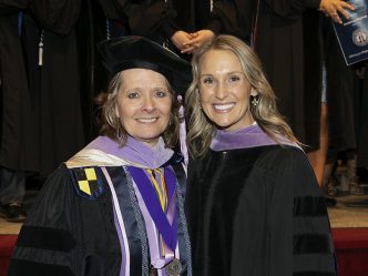 A woman wearing graduation regalia, including doctoral hood, shares a smile with a professor, also in full graduation regalia.