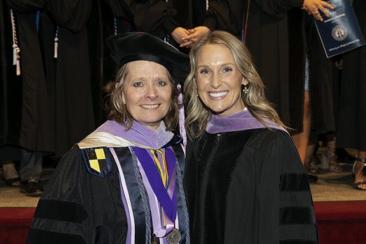 A woman wearing graduation regalia, including doctoral hood, shares a smile with a professor, also in full graduation regalia.