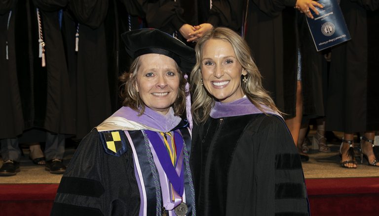 A woman wearing graduation regalia, including doctoral hood, shares a smile with a professor, also in full graduation regalia.