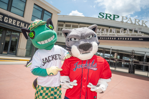 Two mascots, one depicting a green bee in a baseball jersey and another depicting a jaguar in a baseball jersey, stand in front of a minor league baseball stadium.