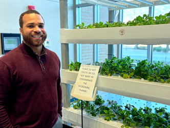 A doctor wearing casual clothes poses beside an indoor micro-farm growing mustard greens.