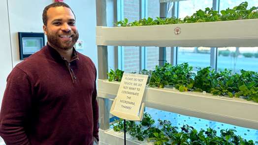 A doctor wearing casual clothes poses beside an indoor micro-farm growing mustard greens.