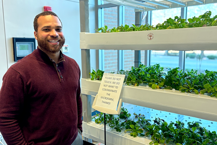 A doctor wearing casual clothes poses beside an indoor micro-farm growing mustard greens.