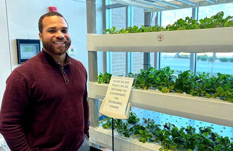 A doctor wearing casual clothes poses beside an indoor micro-farm growing mustard greens.