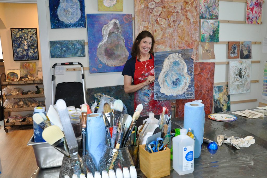 A woman stands in an art studio and holds a painting. She is surrounded by other paintings she has produced.
