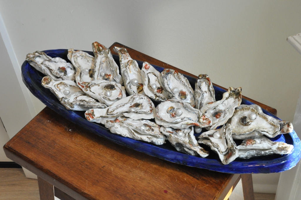 A ceramic tray rests on a table. The tray is lined with open oyster shells with a single pearl resting in each.
