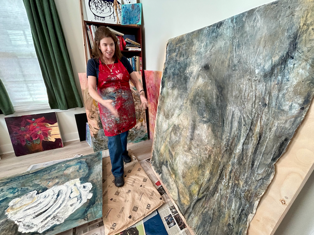 A woman stands in an art studio and gestures toward a painting that is larger than she is. The painting depicts the outside of an oyster shell.