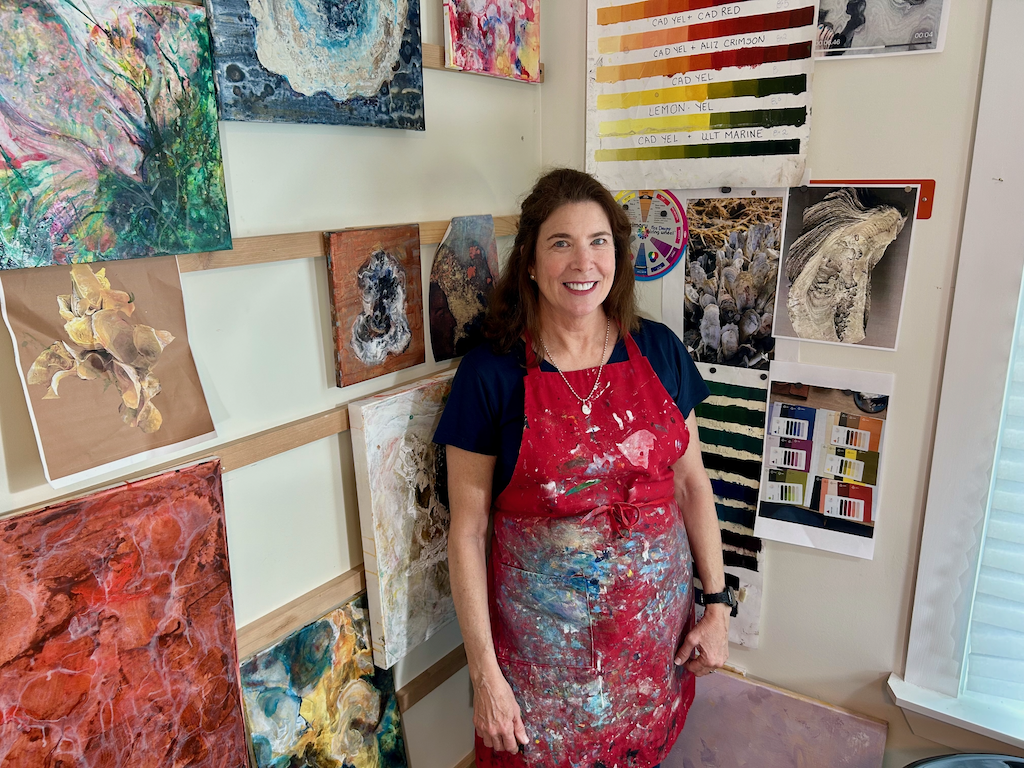 A woman wearing a painting apron stands in front of several of her paintings.