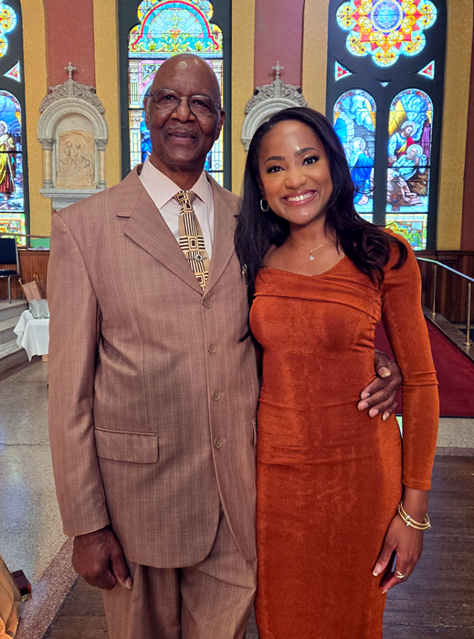 A well dressed man and woman pose for a photo together. Behind them are stained glass windows.