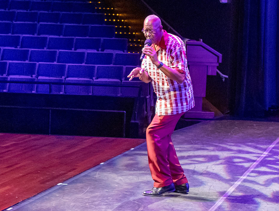 A man wearing dress shoes, slacks and a striped shirt sings into a microphone on stage.