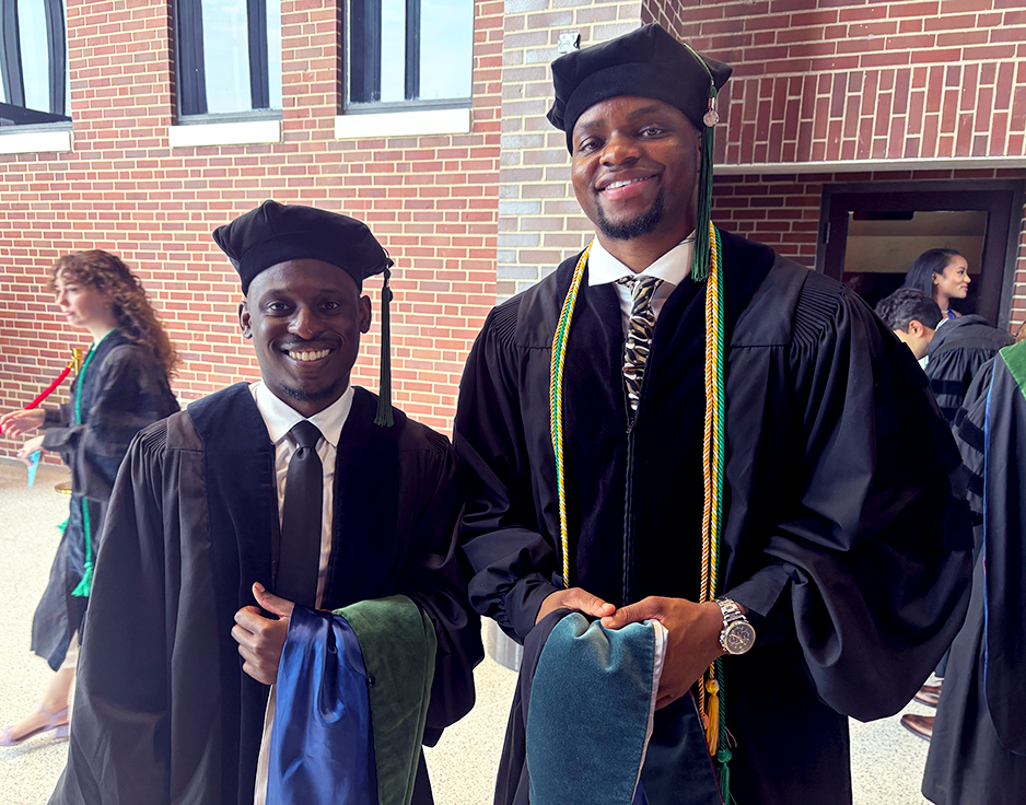 Two men wearing doctorate graduation regalia smile at the camera.