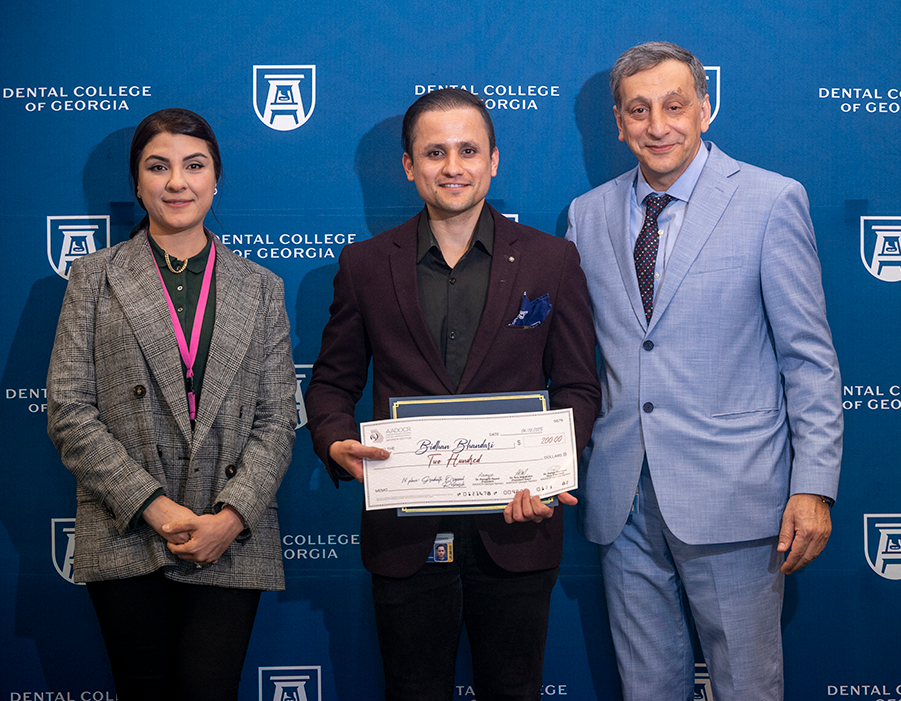 One professionally dressed woman and two professionally dressed men smile in front of a backdrop that reads "Dental College of Georgia." The man in the middle is holding a check aware for two-hundred dollars.