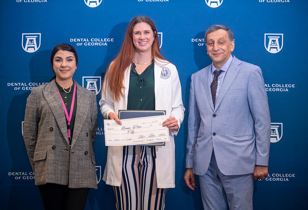 Two professionally dressed women and one professionally dressed man smile in front of a backdrop that reads "Dental College of Georgia." The woman in the middle is holding a check award for fifty dollars.