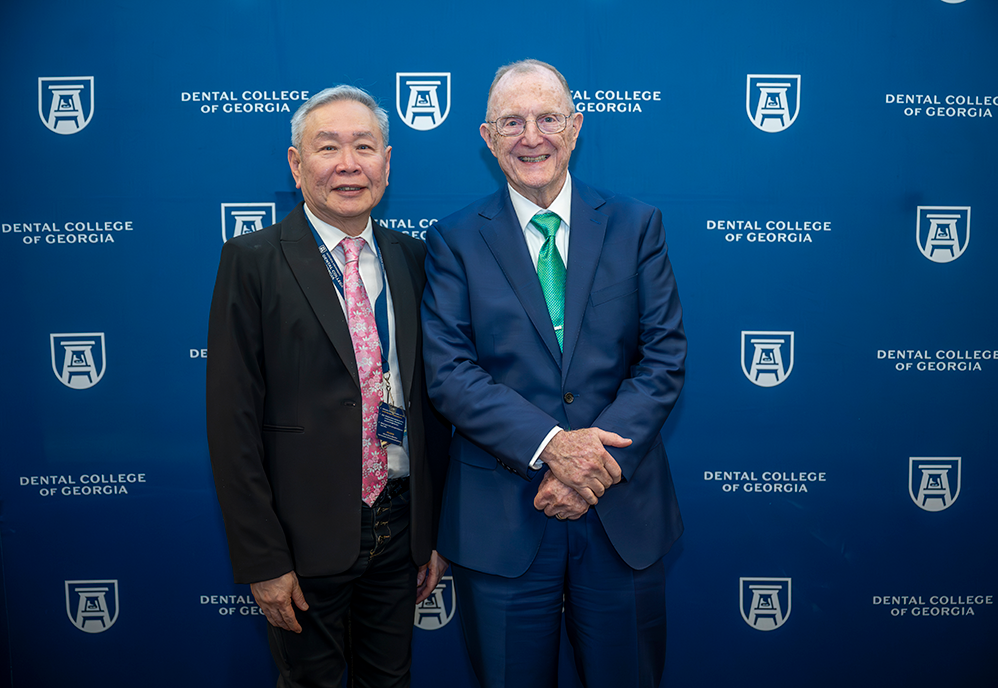 Two professionally dressed men smile in front of a backdrop that reads "Dental College of Georgia."