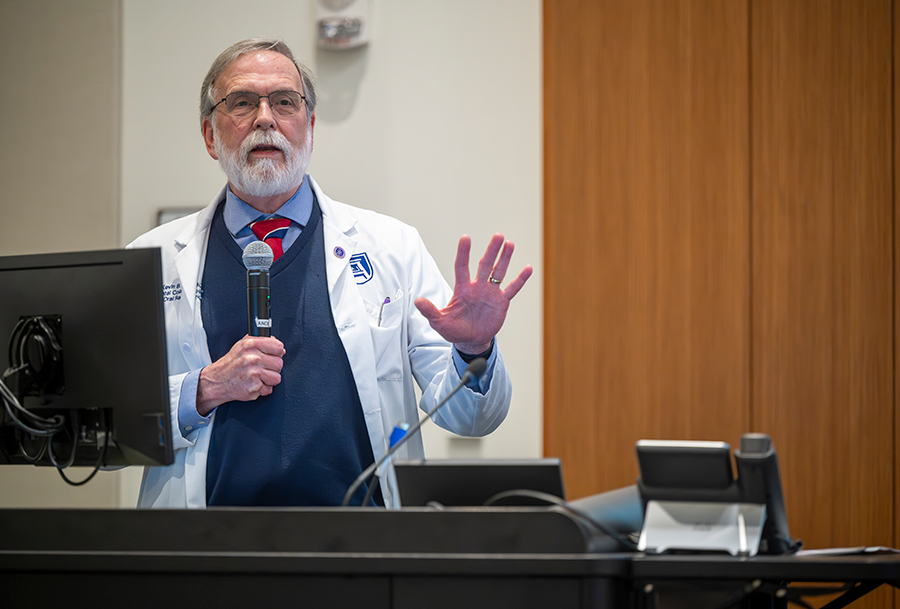 A man wearing a lab coat, holding a microphone speaks at a podium.