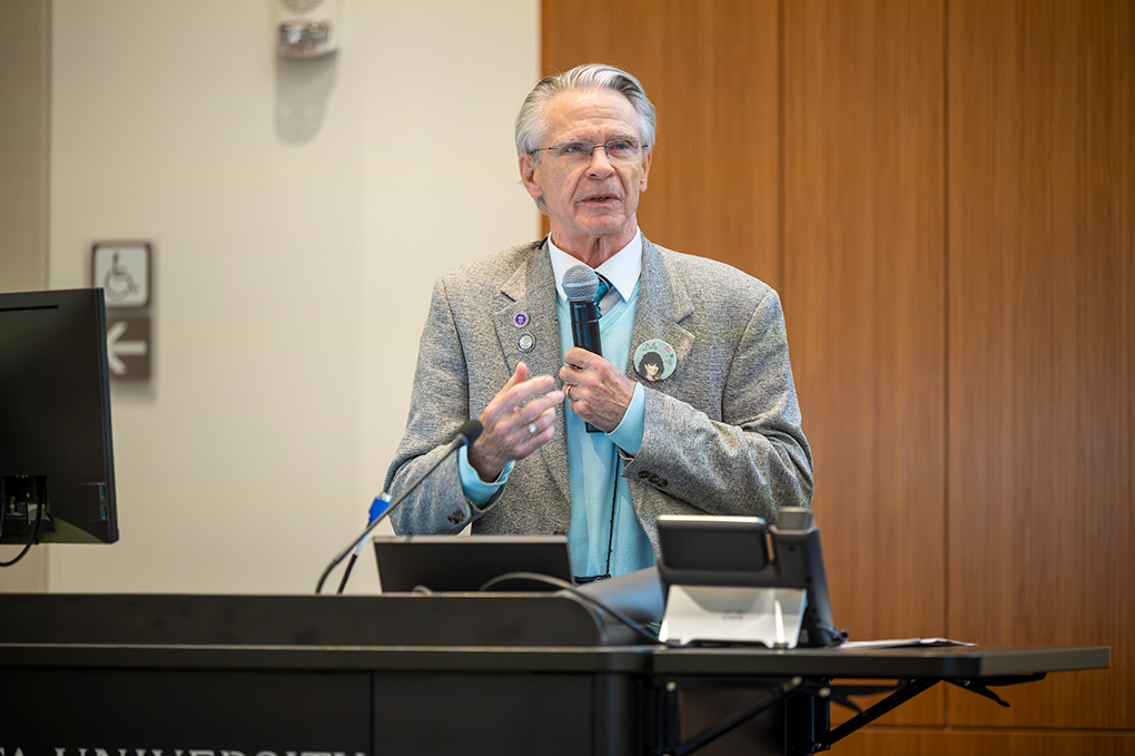 A professionally dressed man holding a microphone speaks at a podium.