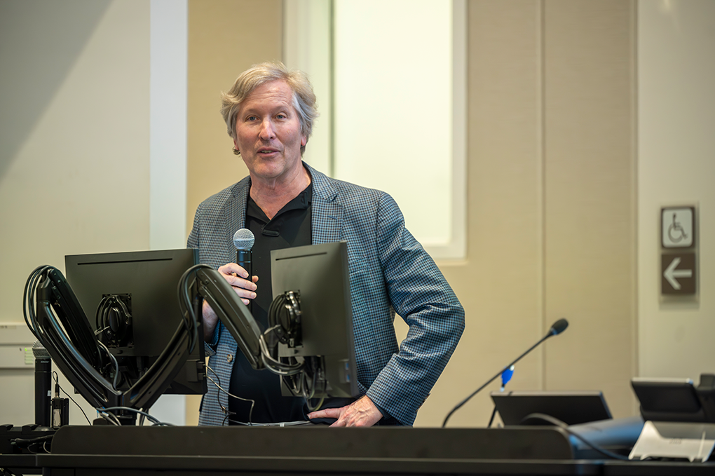 A professionally dressed man holding a microphone speaks at a podium.