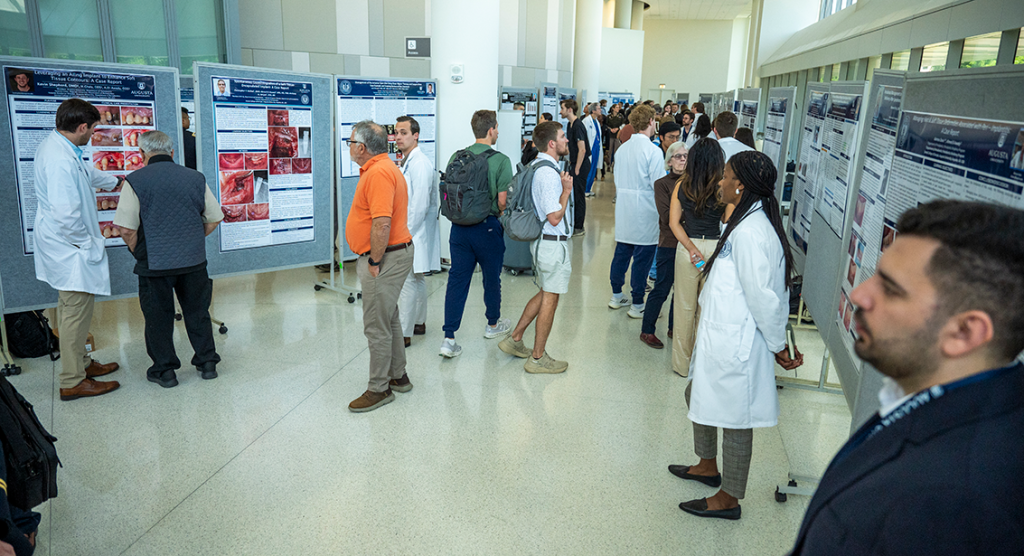 A mixed group of people in lab coats, people with backpacks on and people in casual clothes walk around and observe research posters.