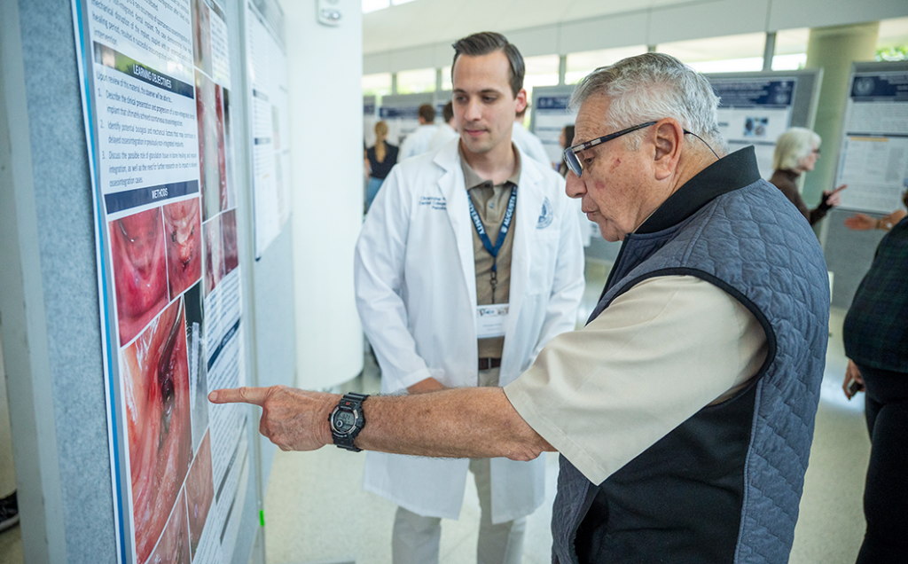 An older man points to a research poster while a younger scientist in a lab coat looks at it.