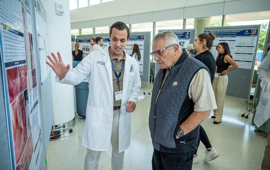 A man wearing a lab coat describes a research poster to a casually dressed man.