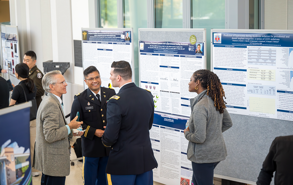 A man and a woman talk to two men wearing military uniforms in front of scientific research posters.