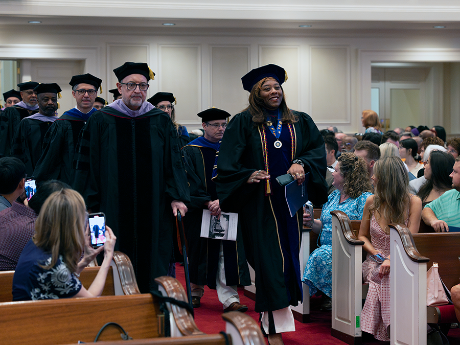 College professors wearing full graduation regalia walk down a center aisle during a graduation ceremony.