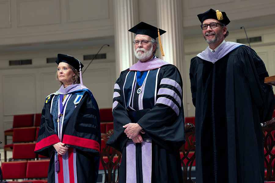 Three college professors, all wearing graduation regalia, stand on a stage during a graduation ceremony.