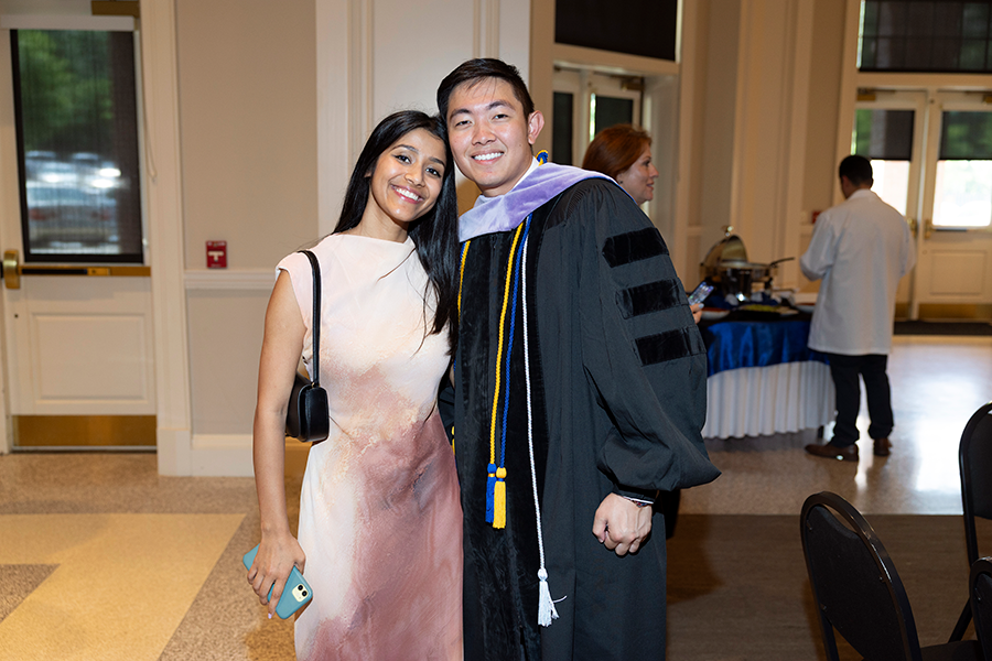 A male college graduate in graduation gown and a ceremonial hood stands with his girlfriend in a large foyer.