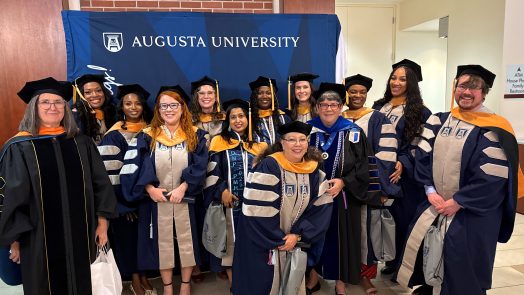 A group of students and faculty in graduation regalia pose together in front of a blue step and repeat