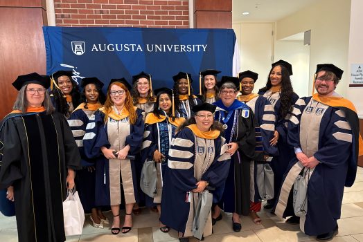 A group of students and faculty in graduation regalia pose together in front of a blue step and repeat