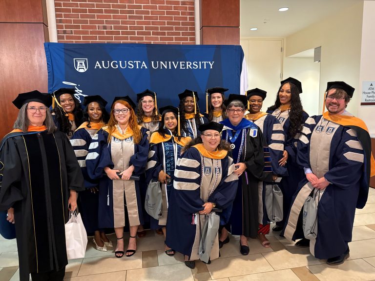 A group of students and faculty in graduation regalia pose together in front of a blue step and repeat