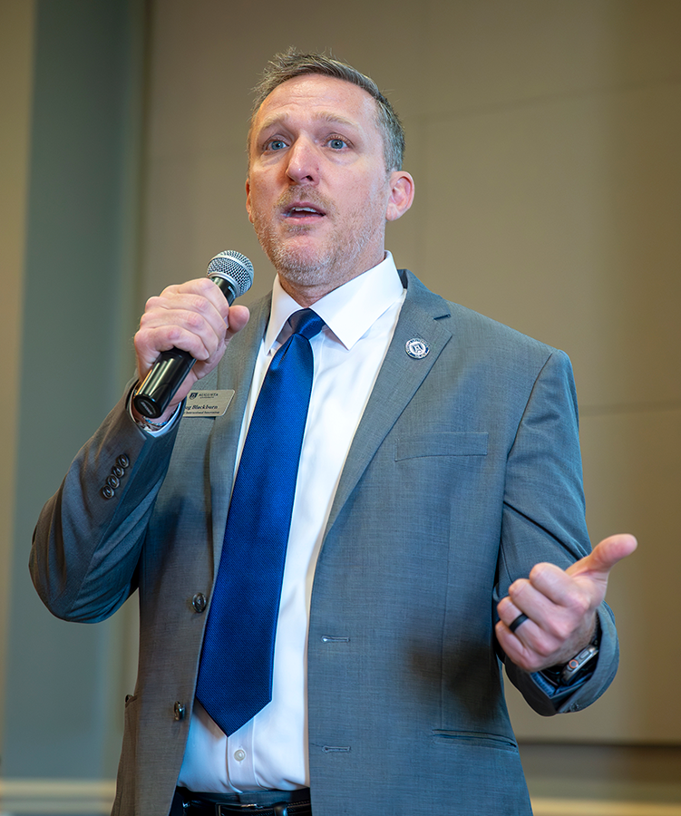 A man holds a microphone and speaks to a group of college faculty during a ceremony in a ballroom.