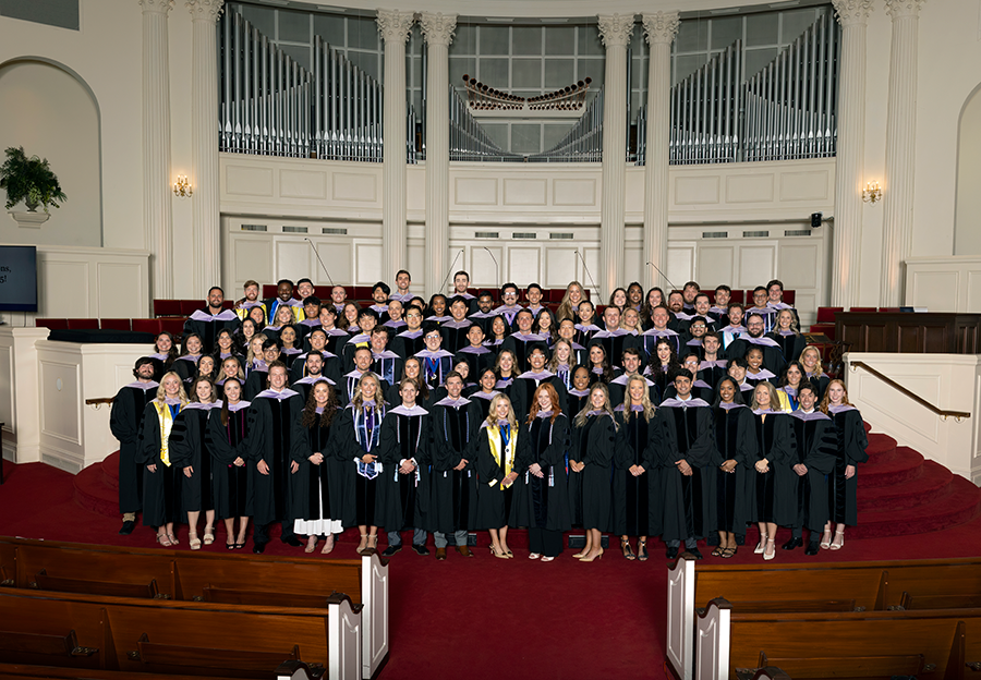 Nearly 100 college graduates, all wearing graduation gowns and ceremonial hoods, stand inside a large church.