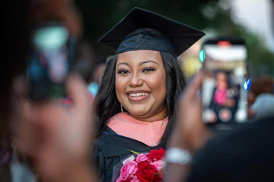 A woman in full college graduation regalia, including a ceremonial hood, stands while holding flowers as her family take photos with their cell phones.