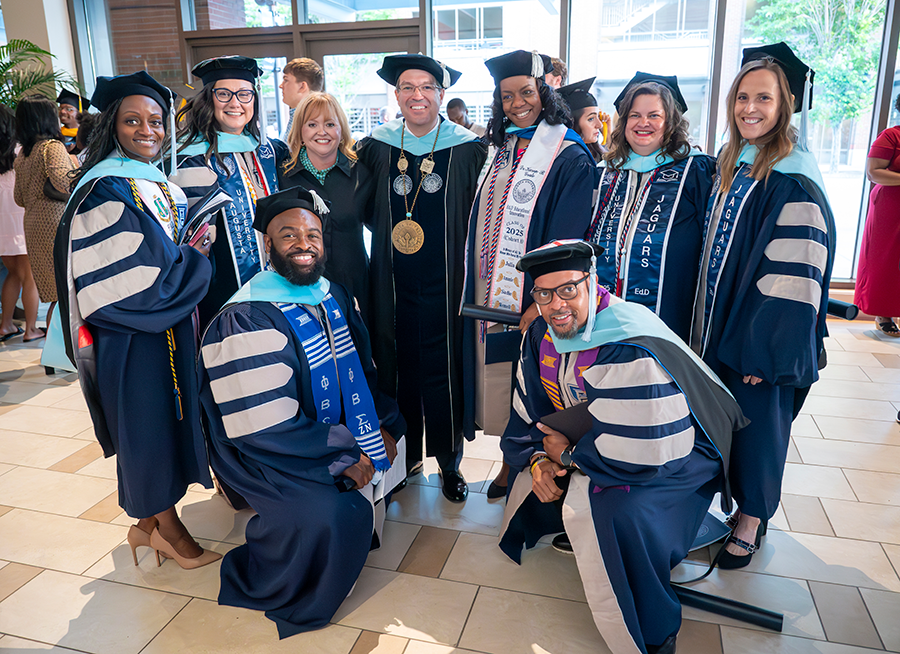 Five women and two men, all wearing full doctoral graduation regalia, pose for a photo with a university president, also in full graduation regalia, and his wife.