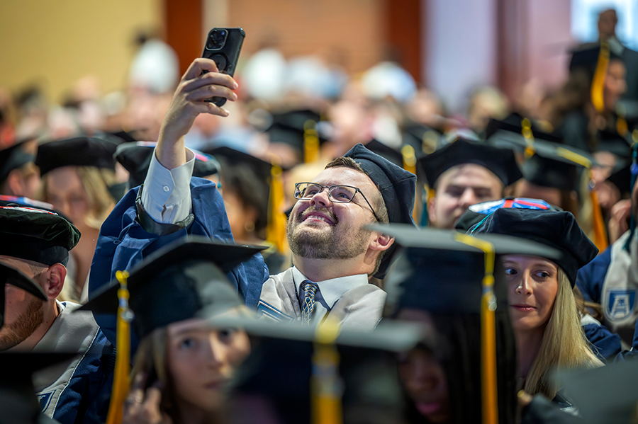 A college male college graduate holds up his cell phone to take a selfie during a college graduation.