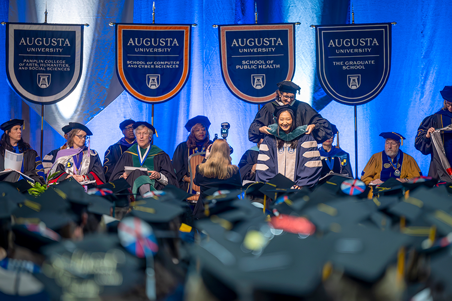 A woman in full college graduation regalia has a ceremonial hood placed around her shoulders during a hooding and commencement ceremony while hundreds of people watch.