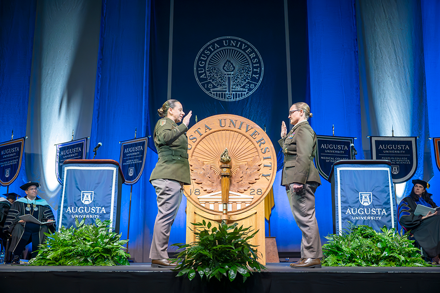 Two women stand on a stage, each holding up their right hands. They are both wearing Army dress uniforms during a special part of a graduation ceremony.