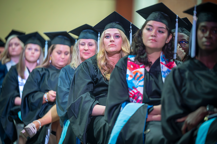 Graduate students in full graduation regalia sit and listen to a guest speaker during a graduation ceremony.