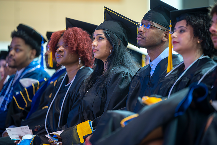 Graduate students in full graduation regalia sit and listen to a guest speaker during a graduation ceremony.