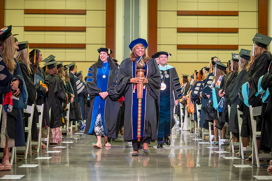 College professors wearing full graduation regalia walk down a center aisle during a graduation ceremony. The female professor in front is carrying a ceremonial mace.