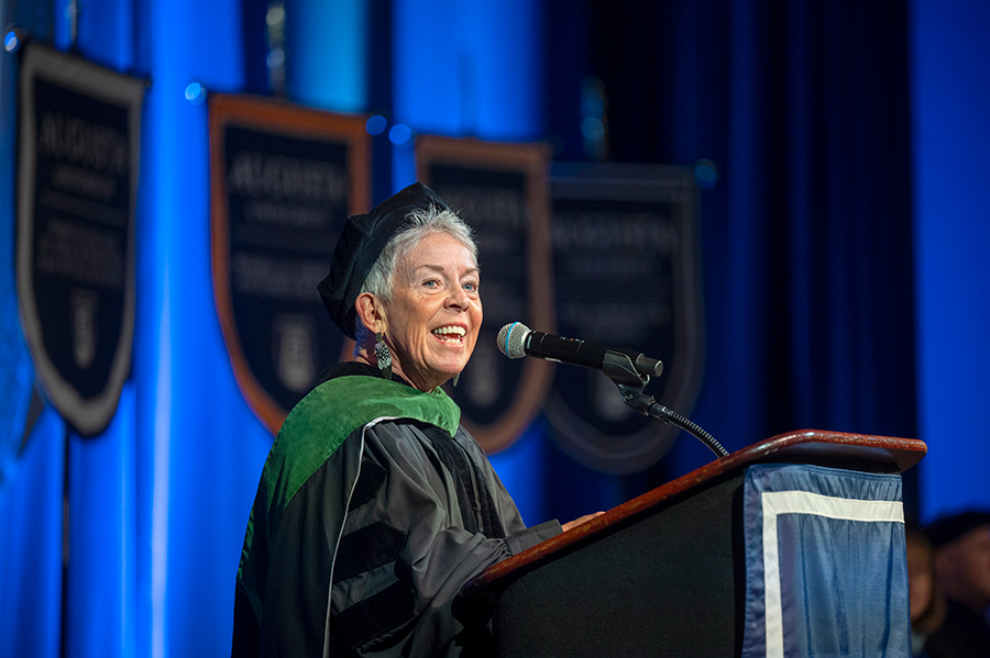 An older woman, dressed in full graduation regalia, stands at a podium with a microphone and delivers a speech to a large group of college graduates.