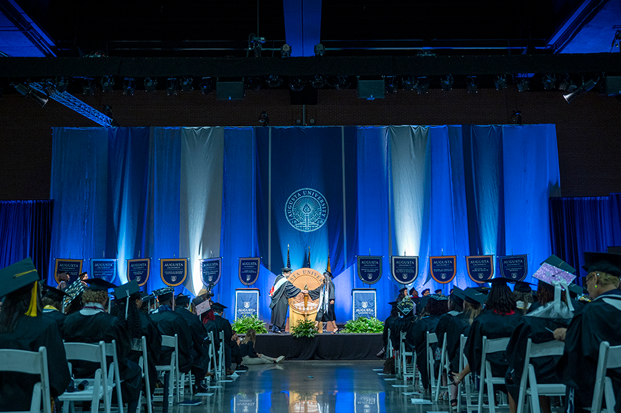 A female college graduate in cap and gown shakes the hand of a male university president during graduation.