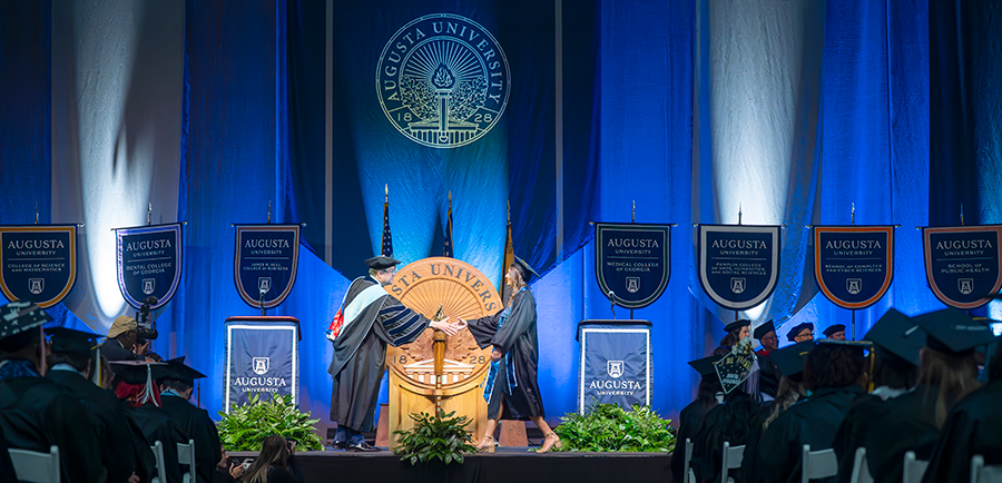 A female college graduate in cap and gown shakes the hand of a male university president during graduation.