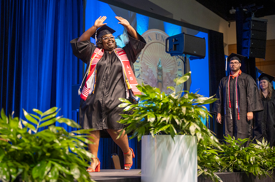 A female college graduate in cap and gown puts her hands up in celebration as she walks across a graduation stage during a large ceremony.