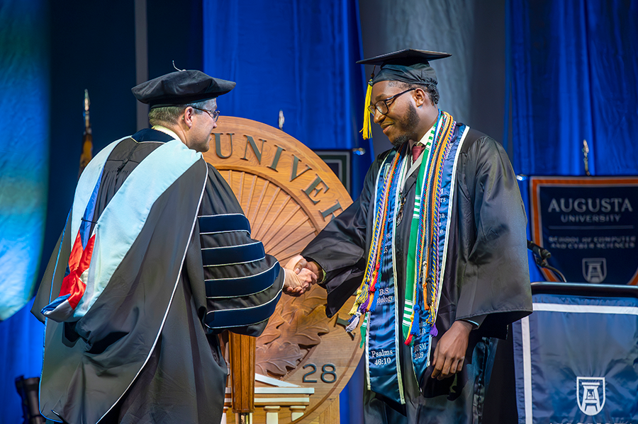 A male college graduate in cap and gown shakes the hand of a male university president during graduation.