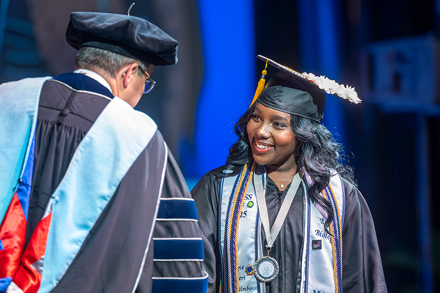 A female college graduate in cap and gown shakes the hand of a male university president during graduation.